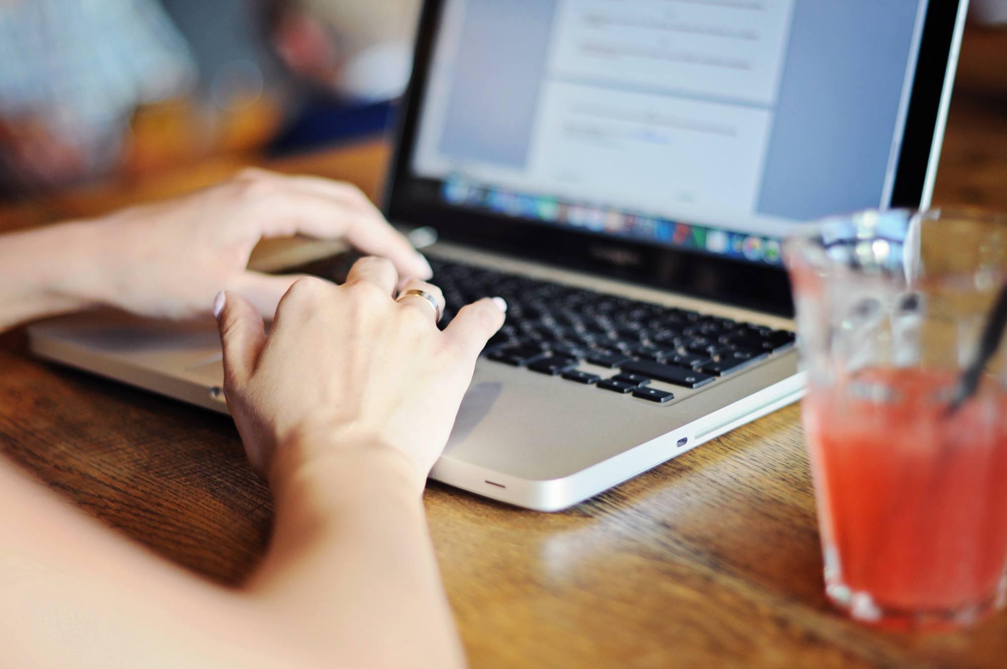 A woman's hands on a laptop with a drink sitting on the table next to it.