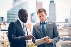 two men in suits looking at a tablet outside with a skyline in the background