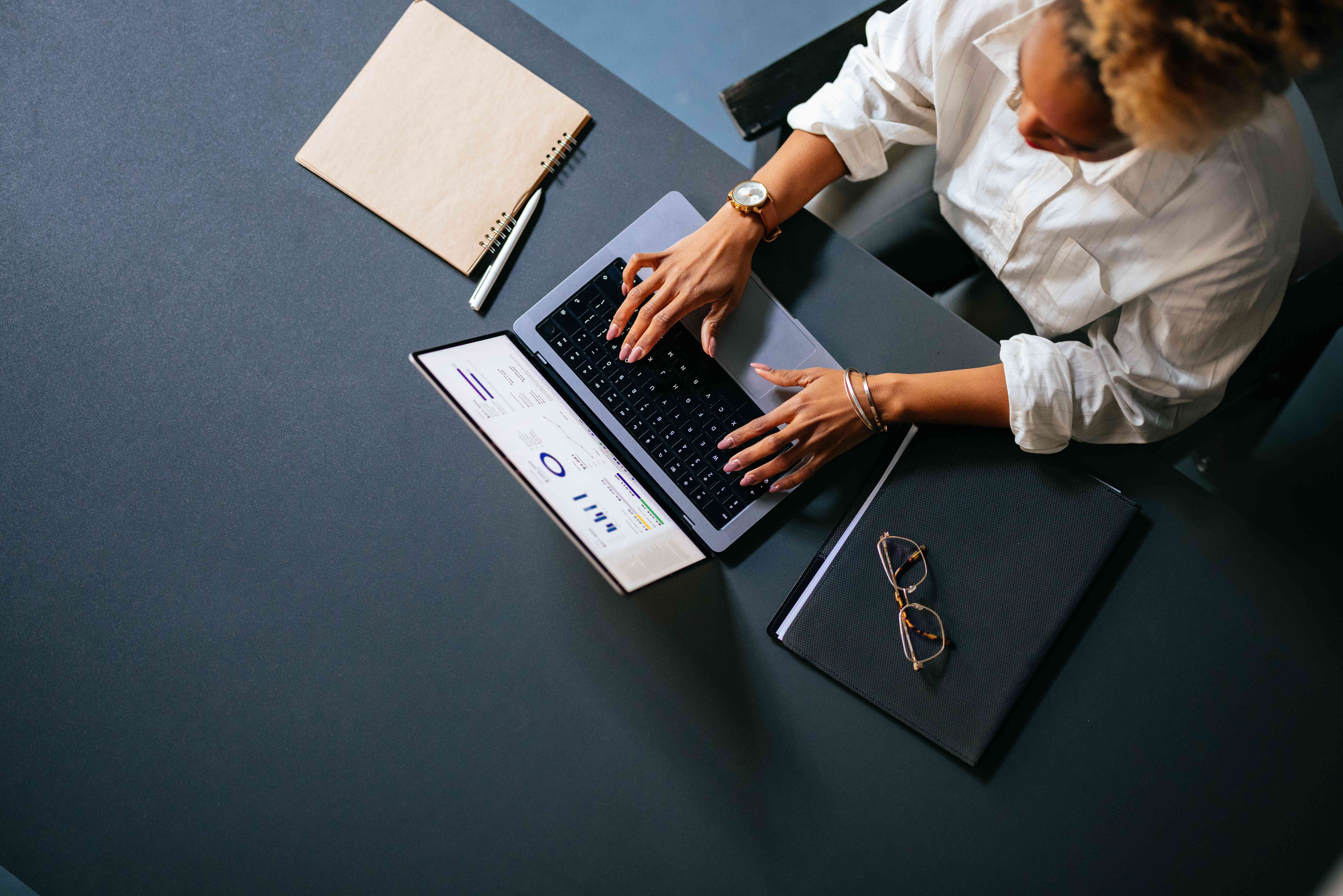 Aerial view of a woman analyzing business graph on a laptop computer while sitting at restaurant desk with notebook, pen, and eyeglasses.