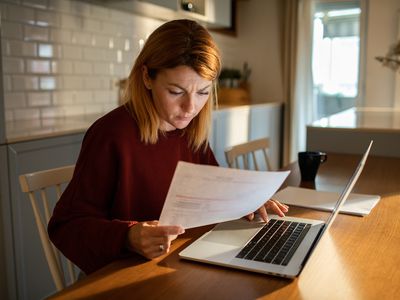 A young woman goes over her finances at a table, with paperwork and a laptop.