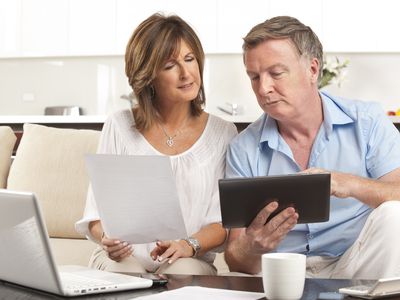 Older couple sitting on living room couch and looking together at a laptop, tablet, and documents