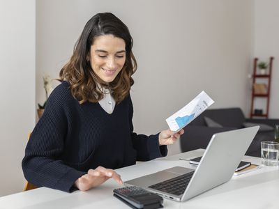 A happy woman uses a calculator and laptop at a desk.