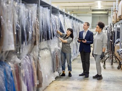 A garment buyer walks down the aisle of a warehouse with two warehouse employees to ensure an order of clothing meets order specifications.