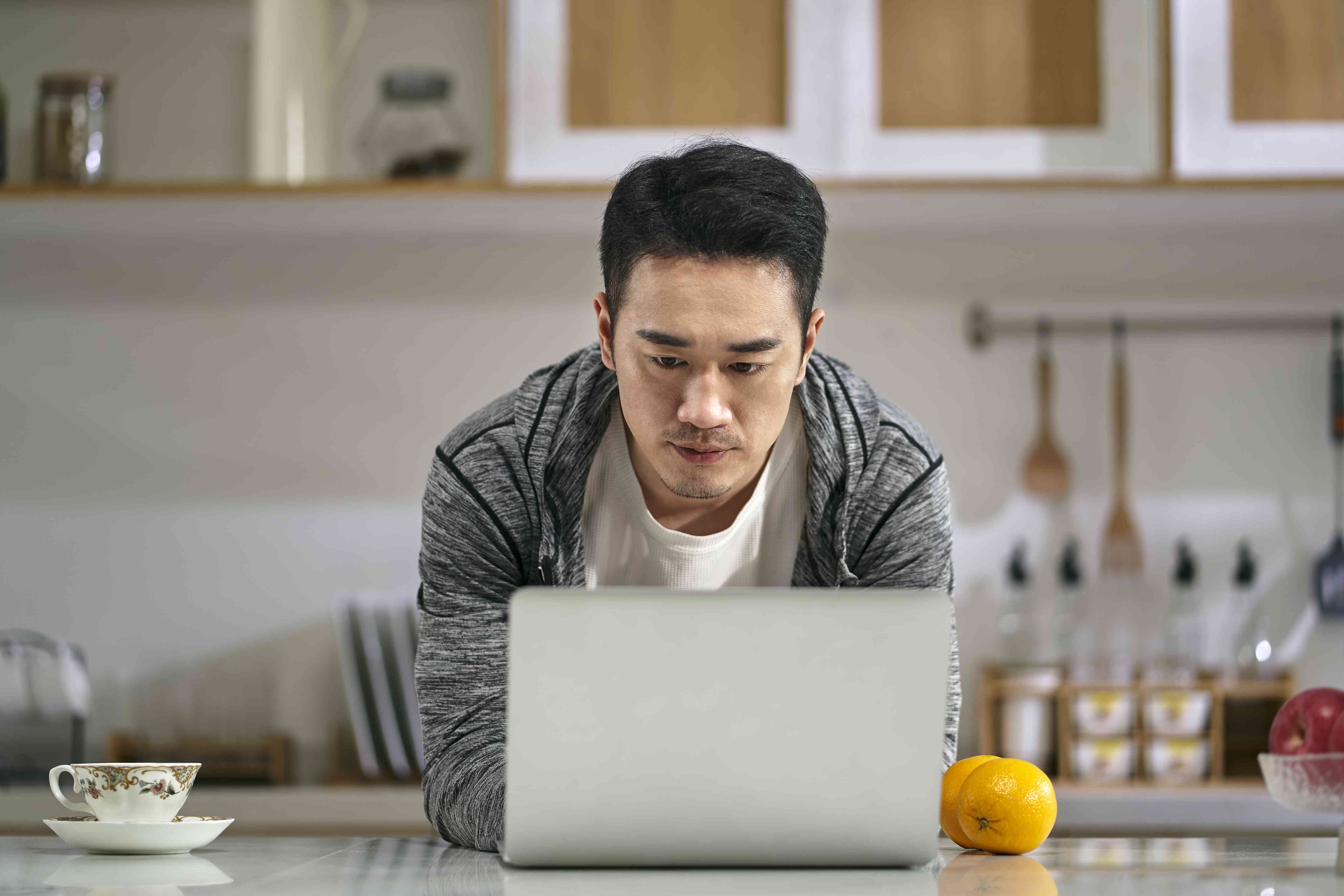 Young man leaning on his kitchen counter as he intently studies something on his laptop