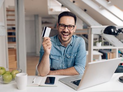 A happy young man sits at a desk with a computer, holding up a debit card.