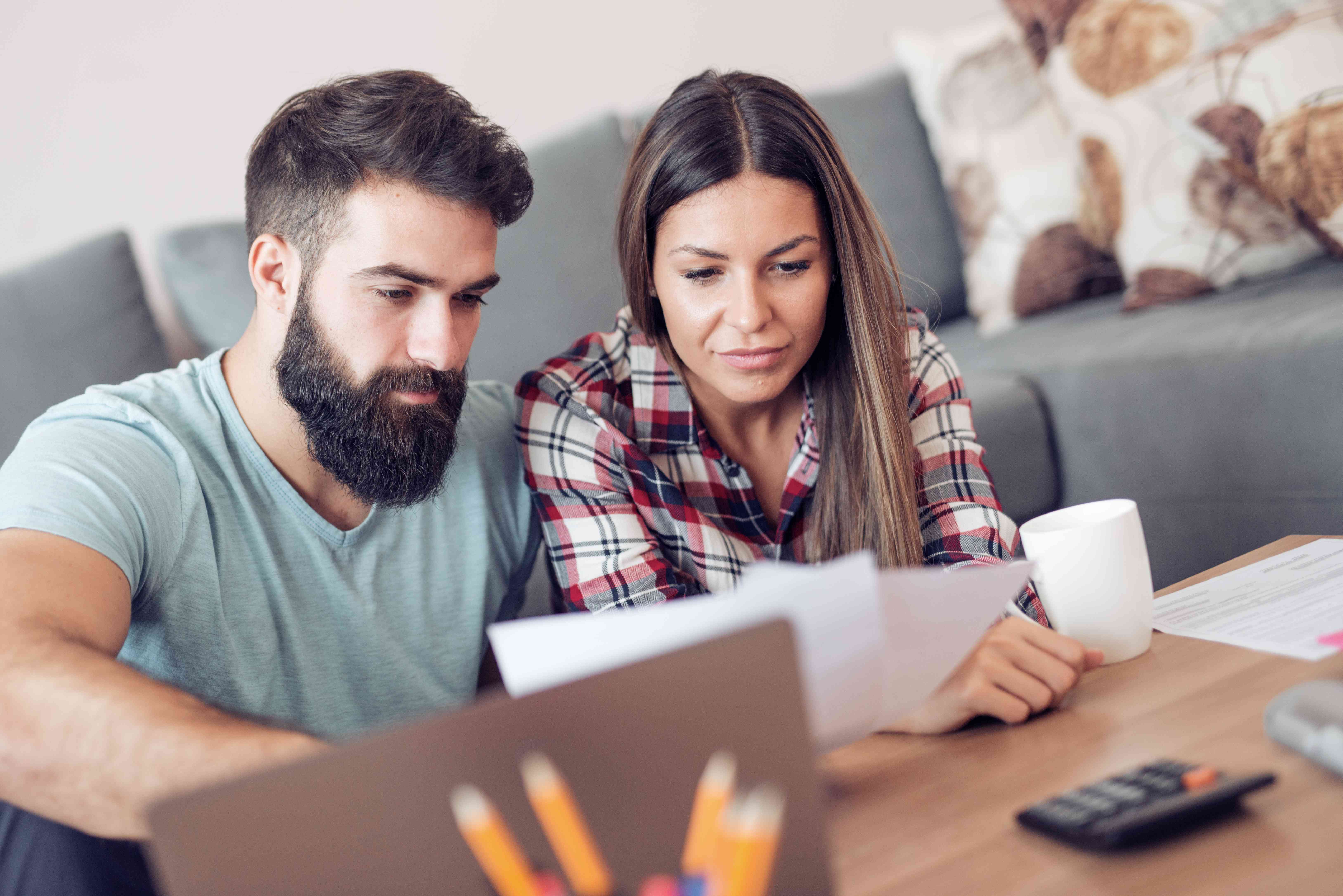 Young couple at home in their living room, looking together at a laptop and financial documents.