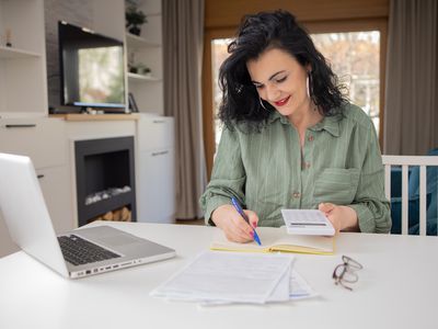 A person sitting in the living room with calculator in one hand and noting down details on notebook about the best options for free checking account with the other hand.