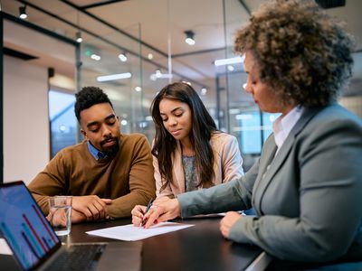 A young couple seated in an office reviews a document as an advisor seated nearby indicates something on the page.