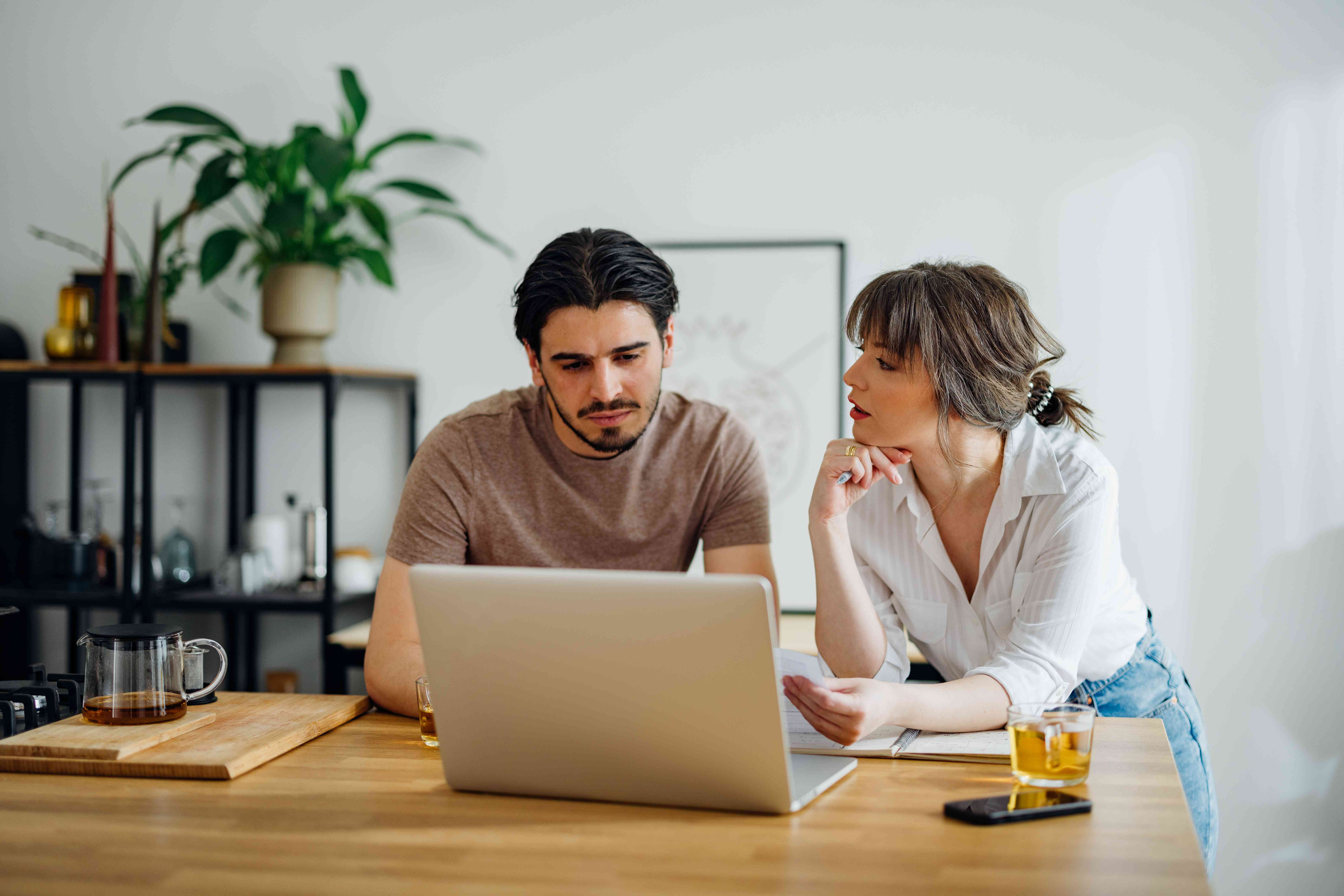 Young couple at their kitchen counter, looking seriously at their laptop and some documents