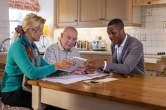 A financial advisor sits at a kitchen table with a senior investor and mature child and explains how they can contribute a gift of funds to an IRA account.