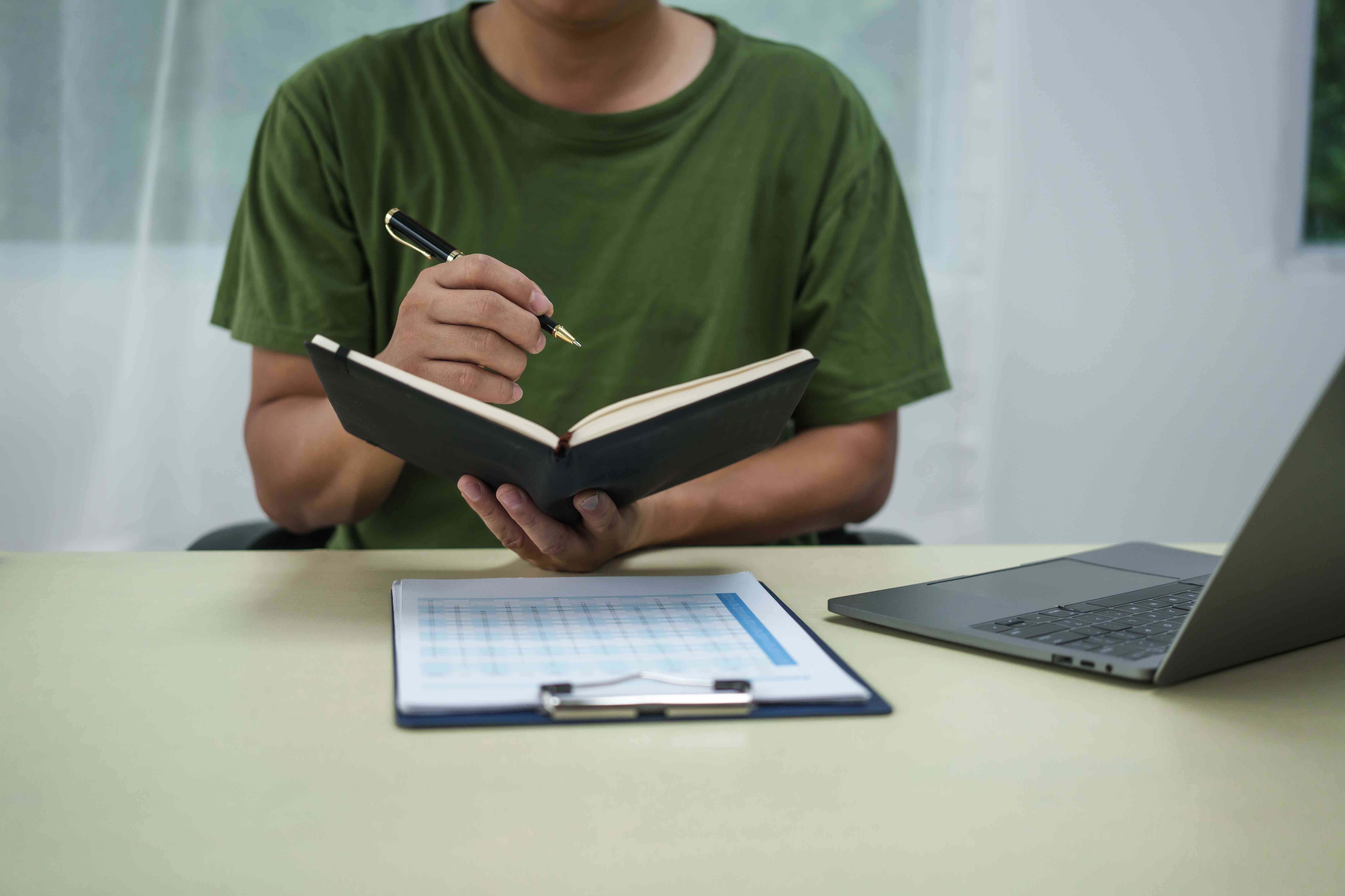 A man writing in an open notebook with an open laptop on the table