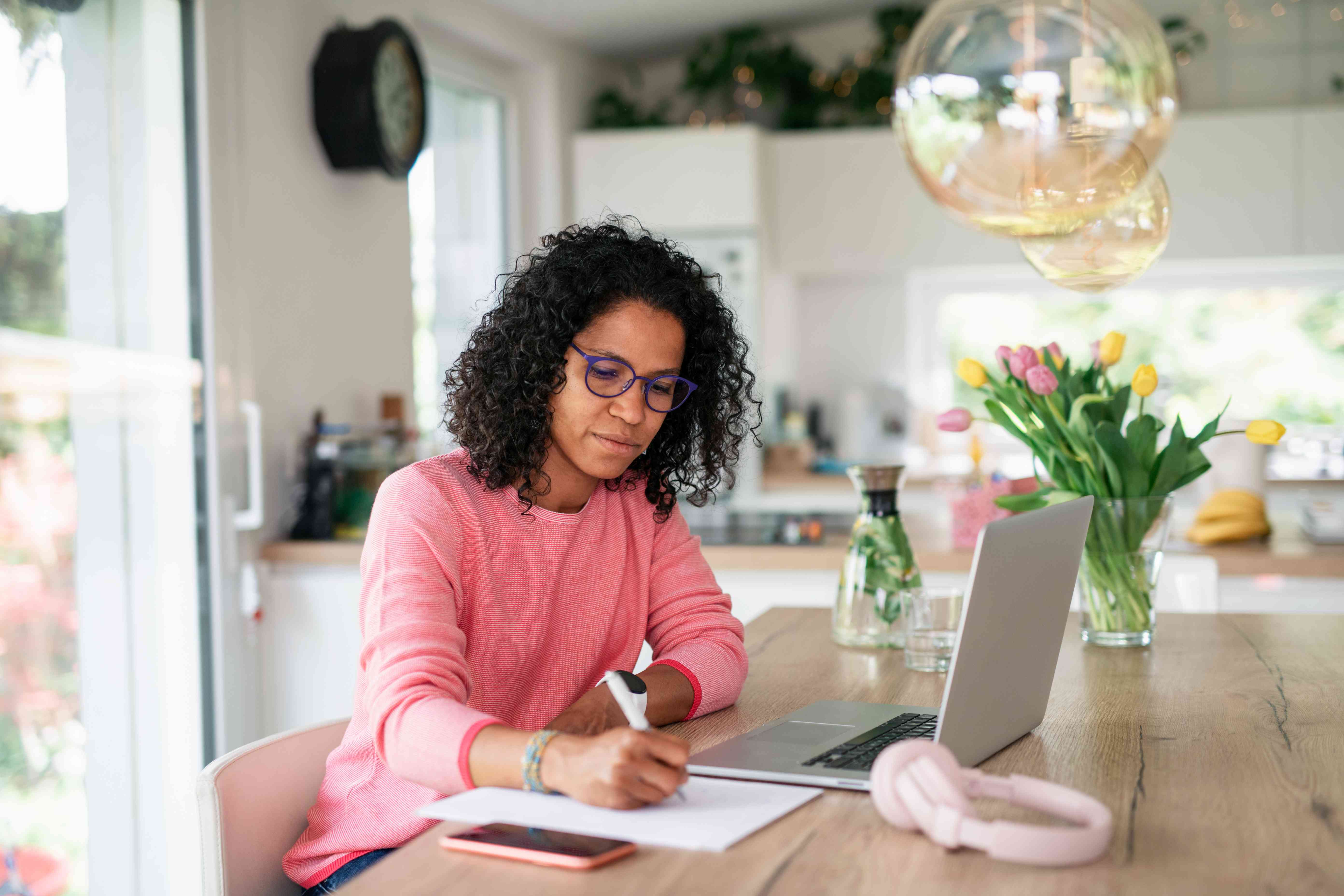 A woman with glasses and a pink shirt sits at a table with a phone, notepad, and laptop computer.