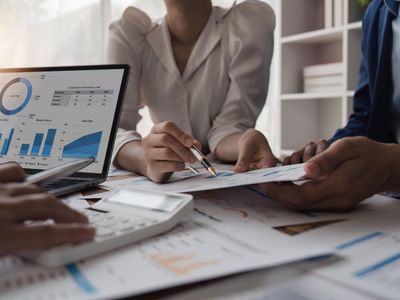 Three mutual fund managers gather around a table covered with financial reports to discuss the performance of various funds the company manages.