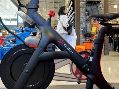 A Peloton bike is displayed in a Dick's Sporting Goods store, with a shopper walking by.