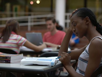 Female student studying with textbook with other students working at desks behind her.