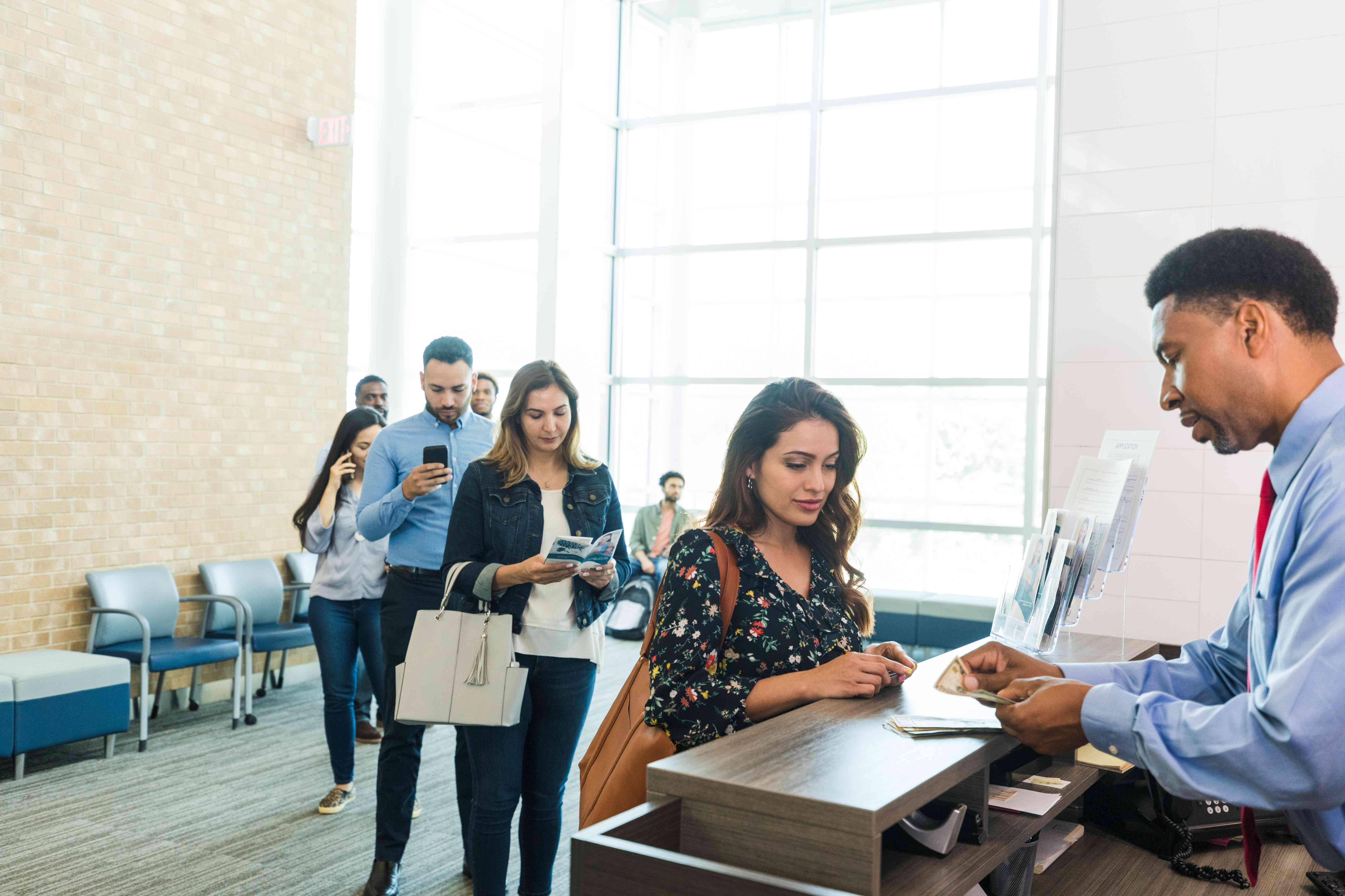 Bank teller counts out cash to a woman at the counter as a line forms behind her. 