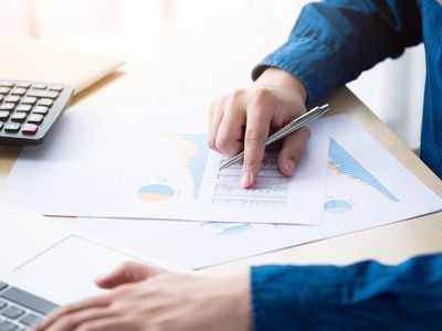 A man types on a laptop with one hand while holding a pen reviewing documents with charts in the other hand.