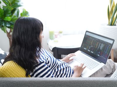 A woman sits on a couch with a laptop in her lap, looking at financial data on the computer.