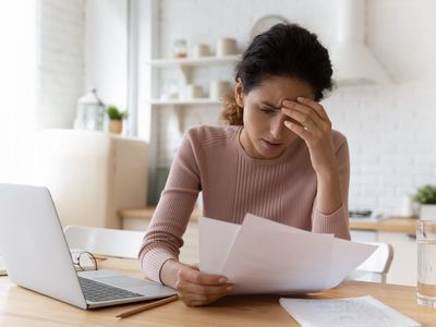 A worried woman looks at paper bills at a table, with a laptop computer.