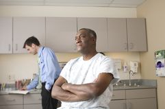 Black man with arms crossed in patient gown while White doctor writes in file