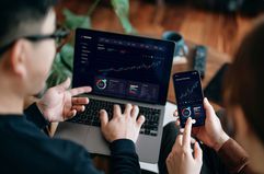 Two people looking at market charts on a laptop and smartphone