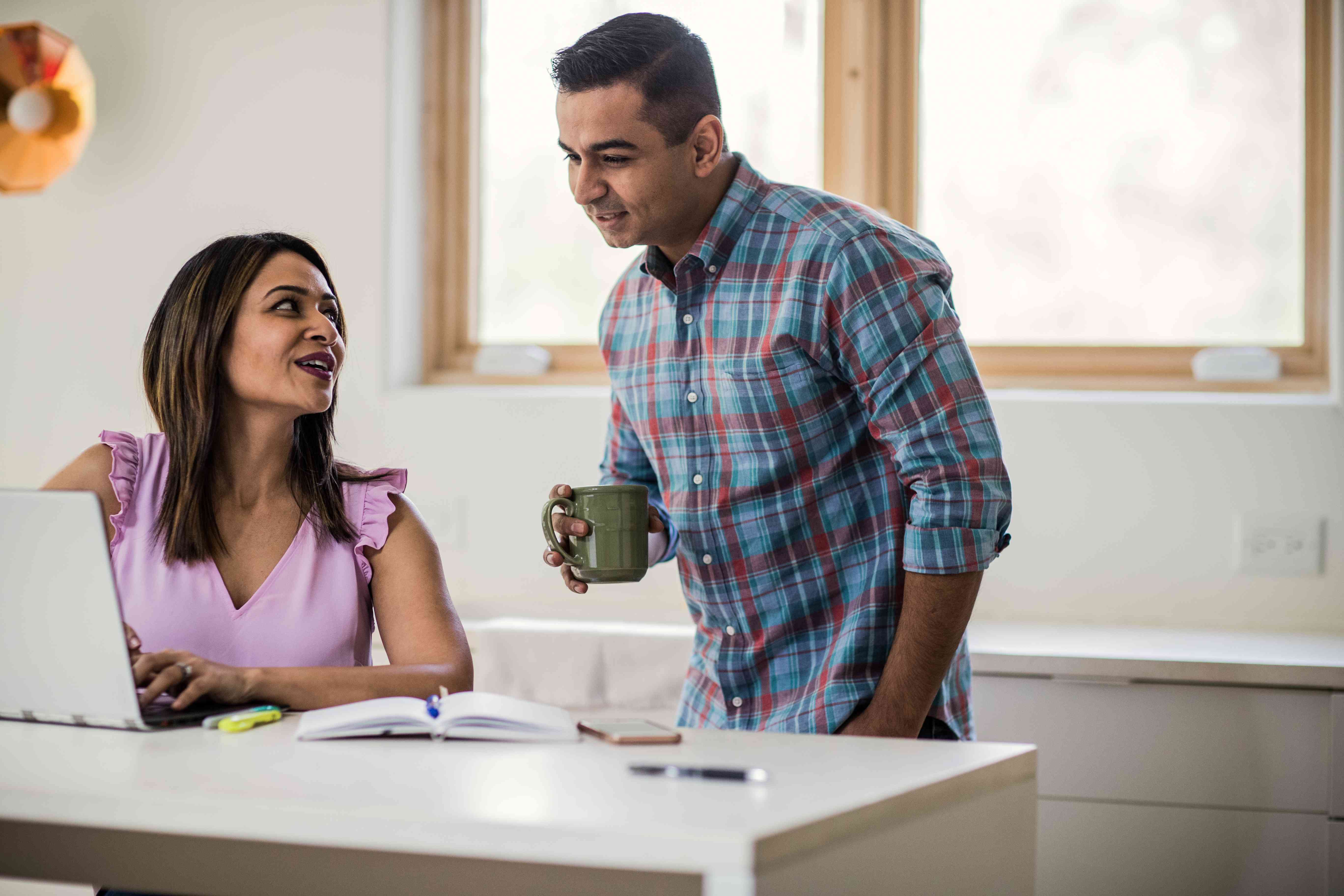 A man holds a coffee cup while chatting with a woman who is sitting with a laptop at a counter