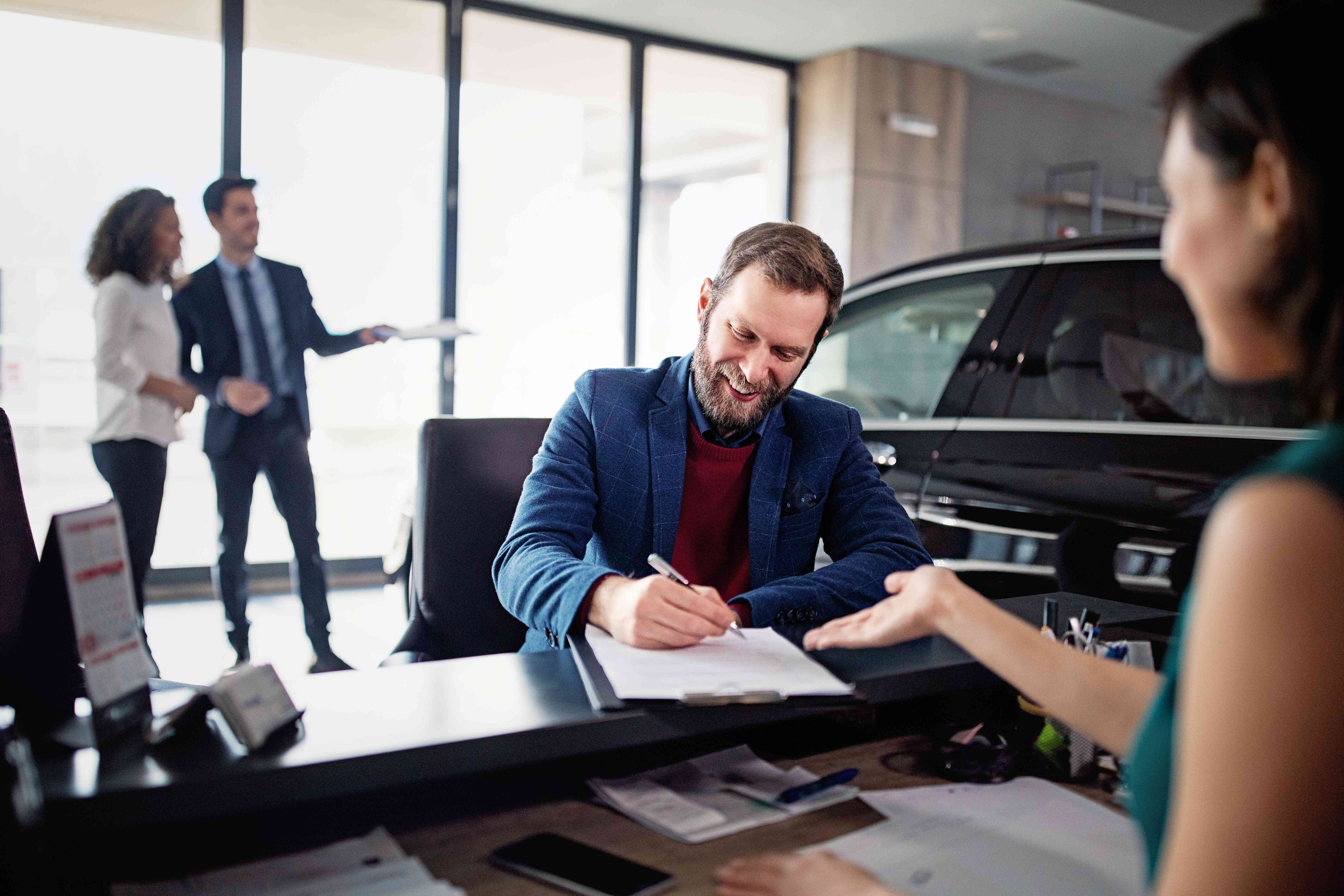 A man signs documents at a car dealership desk to buy a car.