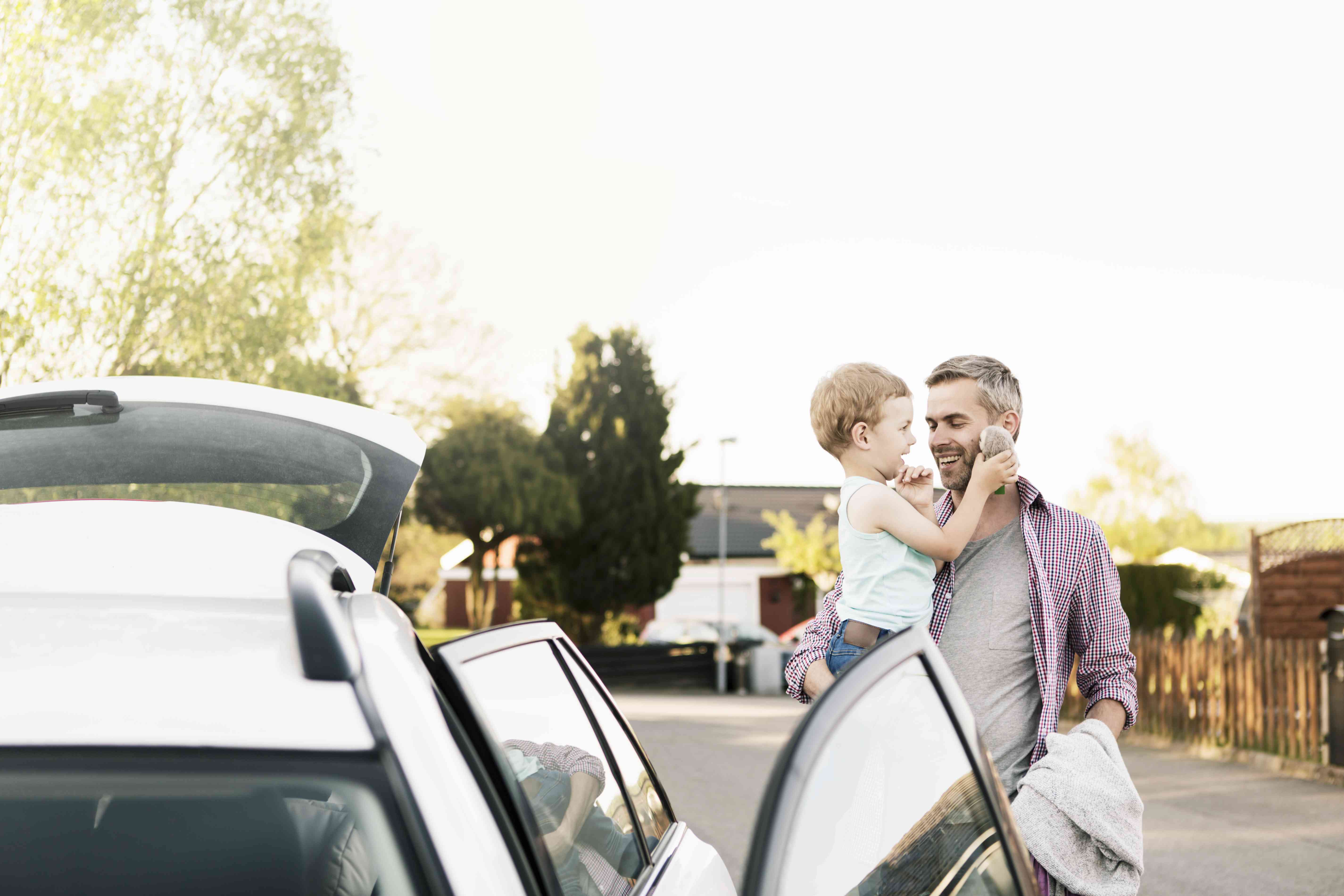 A man stands by a car and holds a toddler.