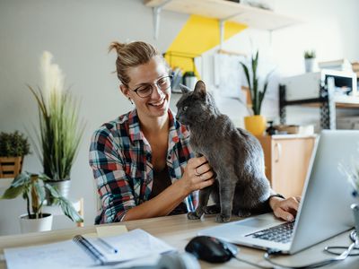 Woman working on laptop computer in a home office while petting her cat