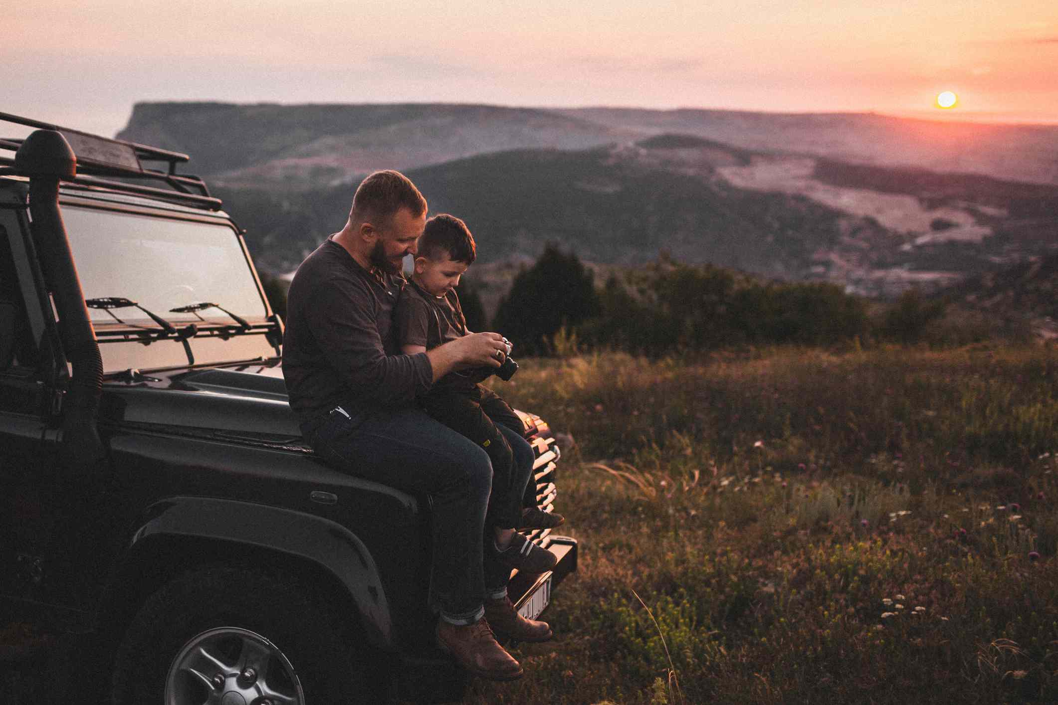 Father and son sitting on the hood of a car parked in nature and looking at a camera