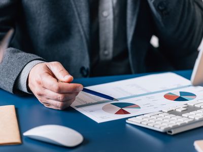 A man wearing a suit sits at a desk and points with a pen to a paper with financial charts.