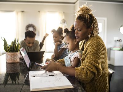 Mother working from home while holding toddler, family in background