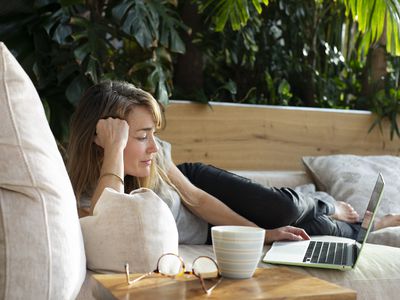 A woman lays on a couch with a laptop, mug, and glasses