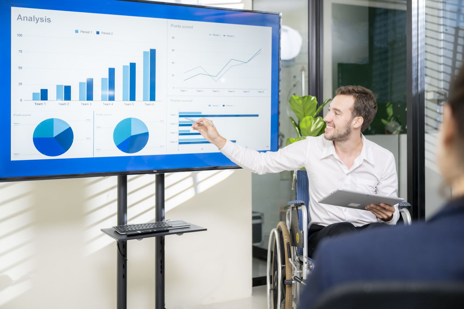 An executive who uses a wheelchair gives a presentation using a large projection chart showing the company's profits.