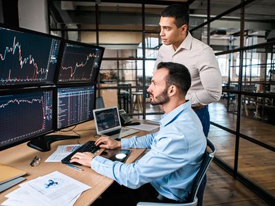 Two traders, one sitting, the other standing looking at several computer monitors displaying financial data.