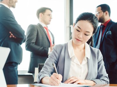 Seated woman works at a table while male colleagues chat in background