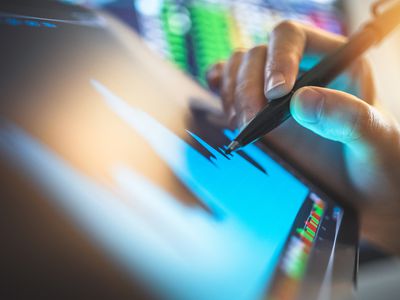 a hand holding a stylus used on a tablet with a chart with stock tickers in the background