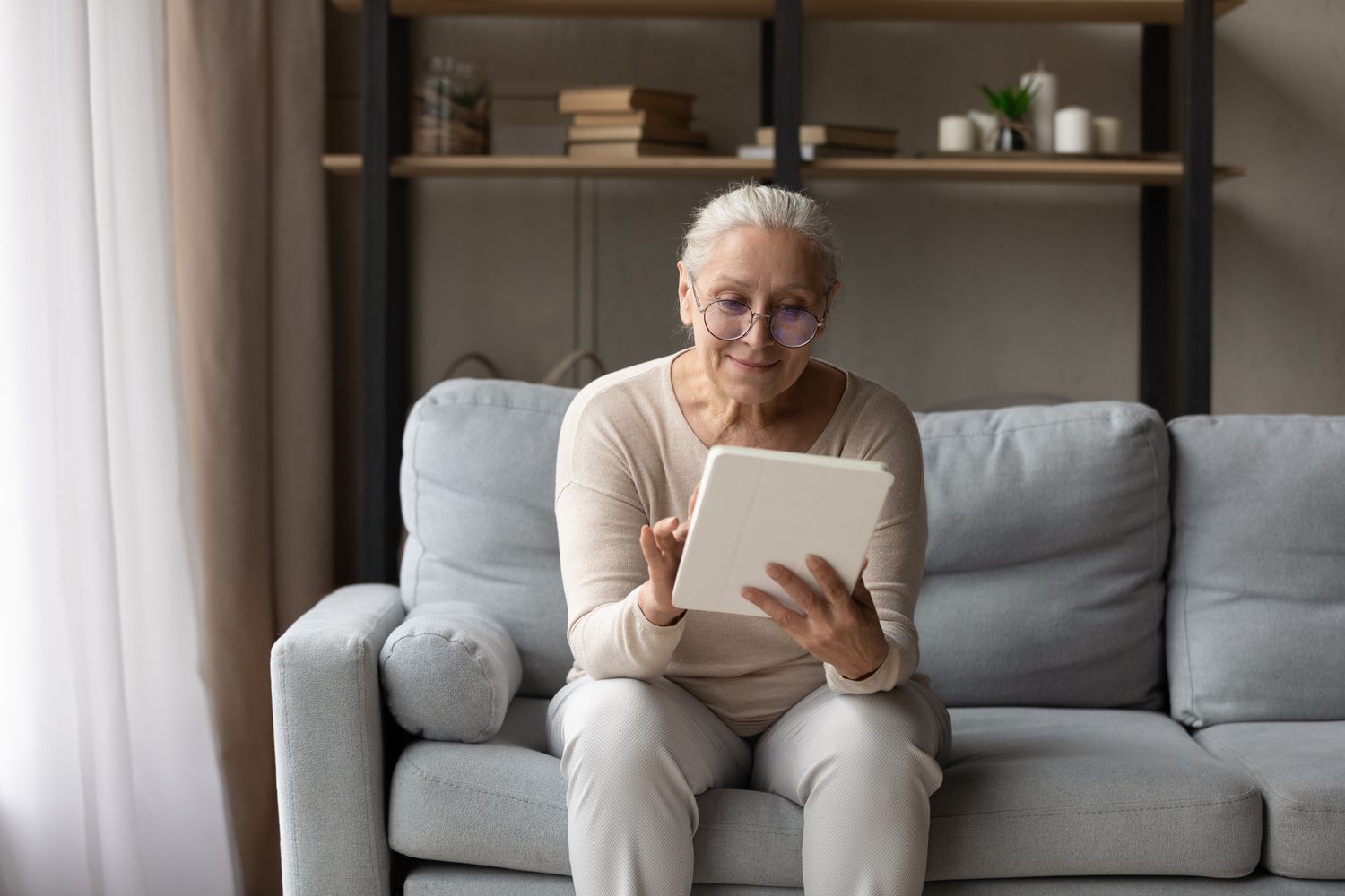 Older woman sitting on her living room couch and smiling as she broswes on her tablet