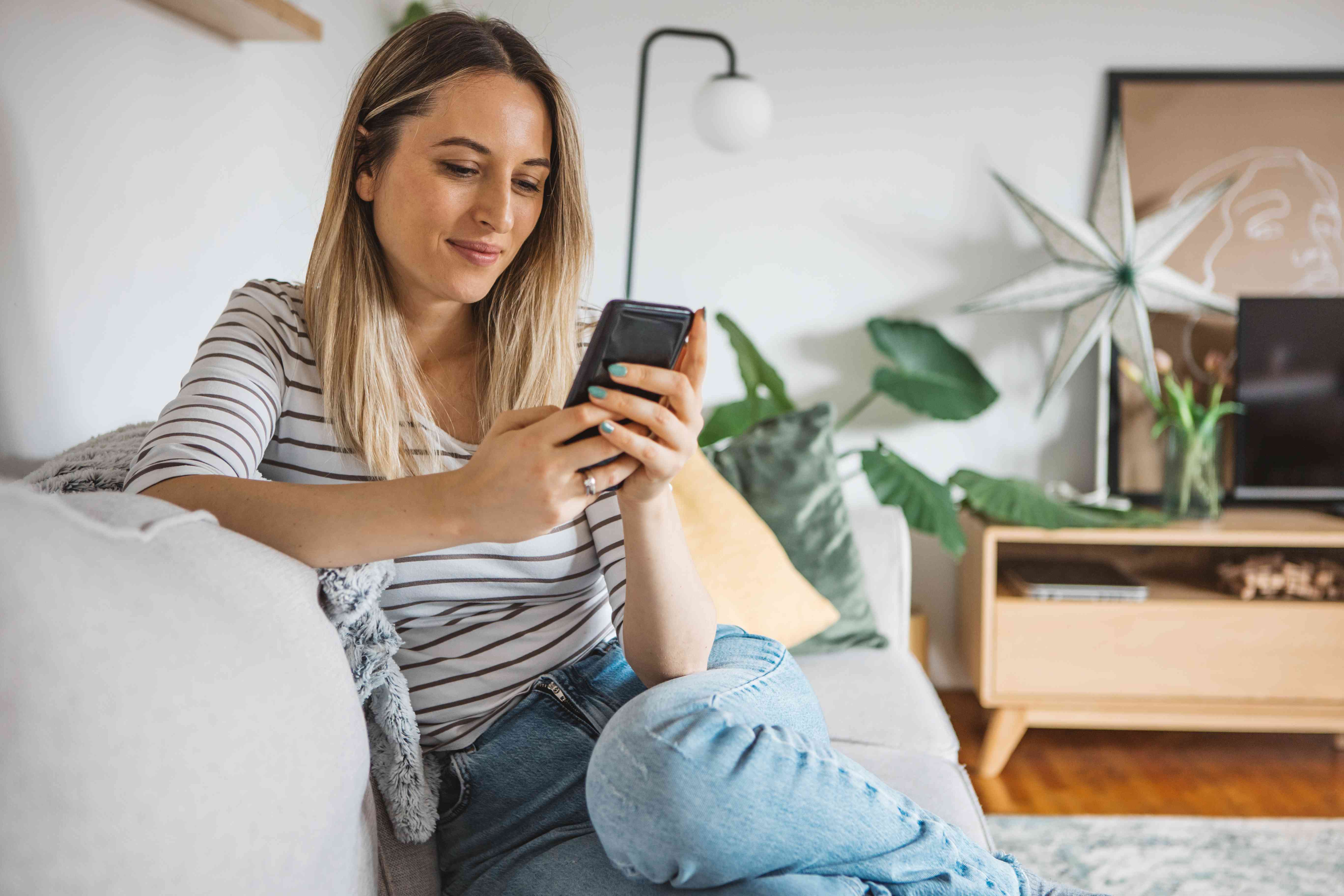 Yougn woman sitting on her living room couch and smiling as she looks at her smartphone