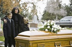 A widow stands with their arms around two young children next to a casket at a cemetery.