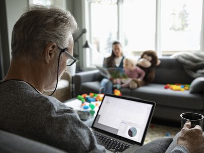 A retiree sits in an overstuffed chair with a mug of coffee reviewing their annuity account on a laptop while their grandchild reads a book with the grandmother on a sofa across the room.