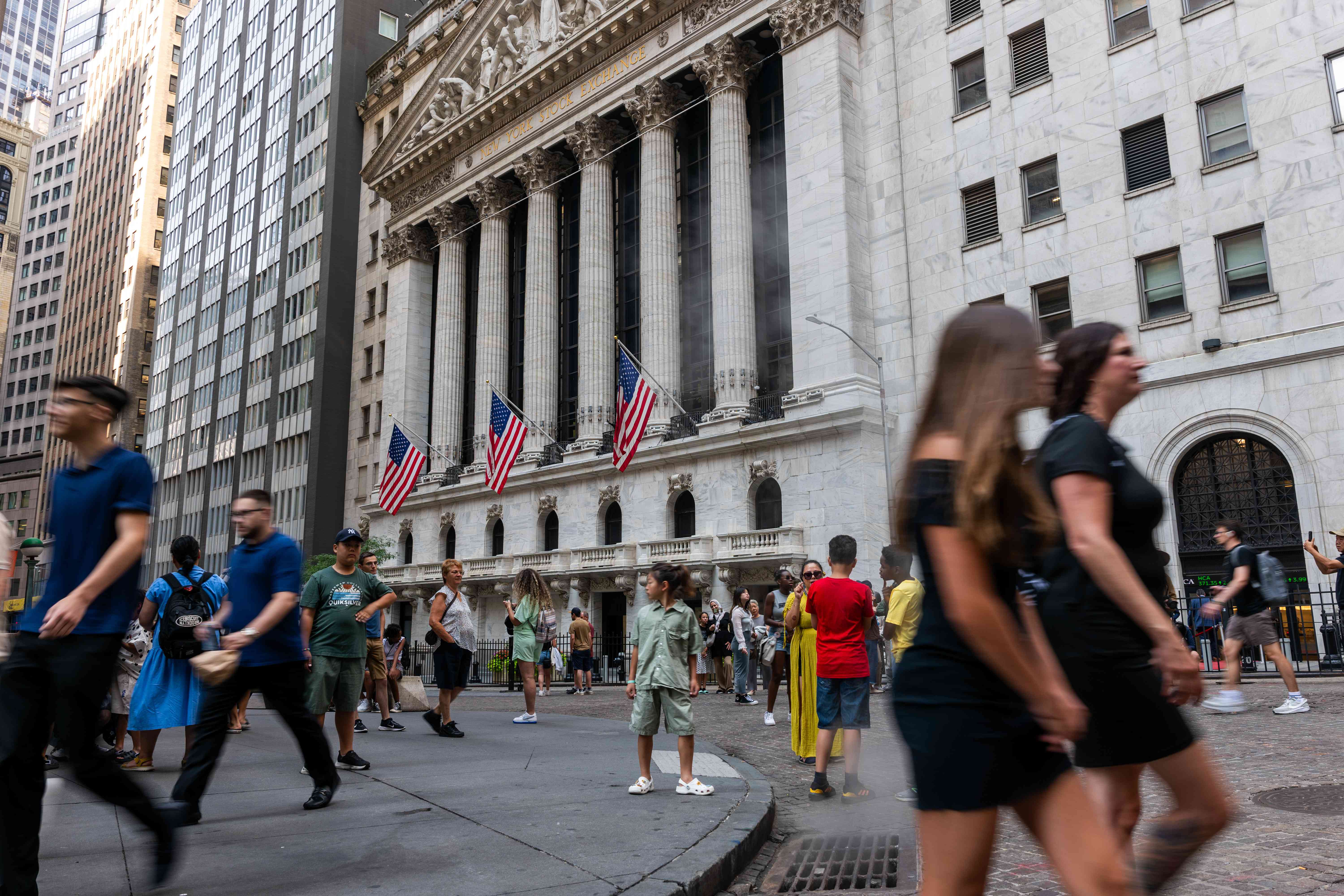 Several people walk past outside the New York Stock Exchange building, where three American flags are on display.