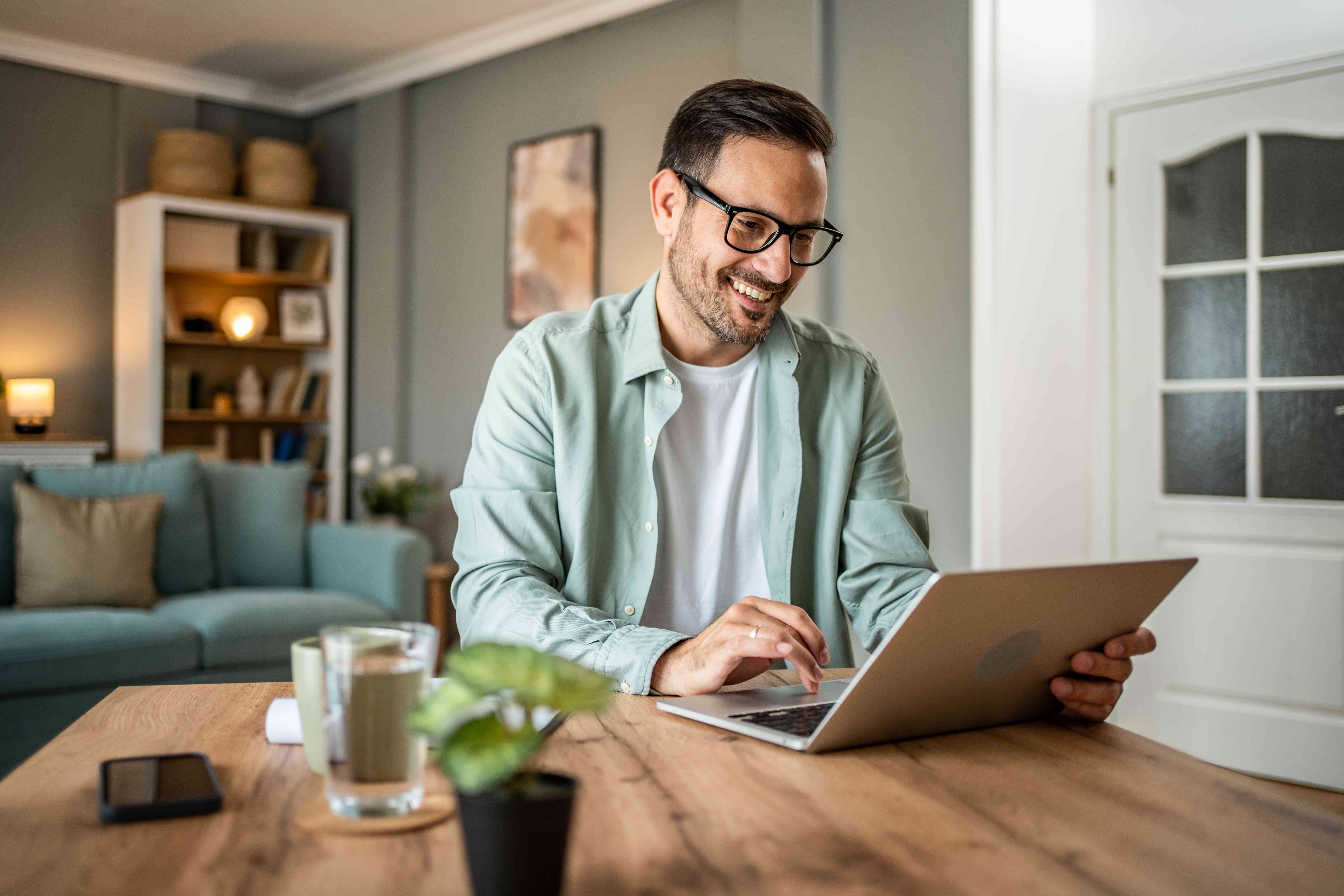 A smiling man uses a laptop while sitting at a table at home.