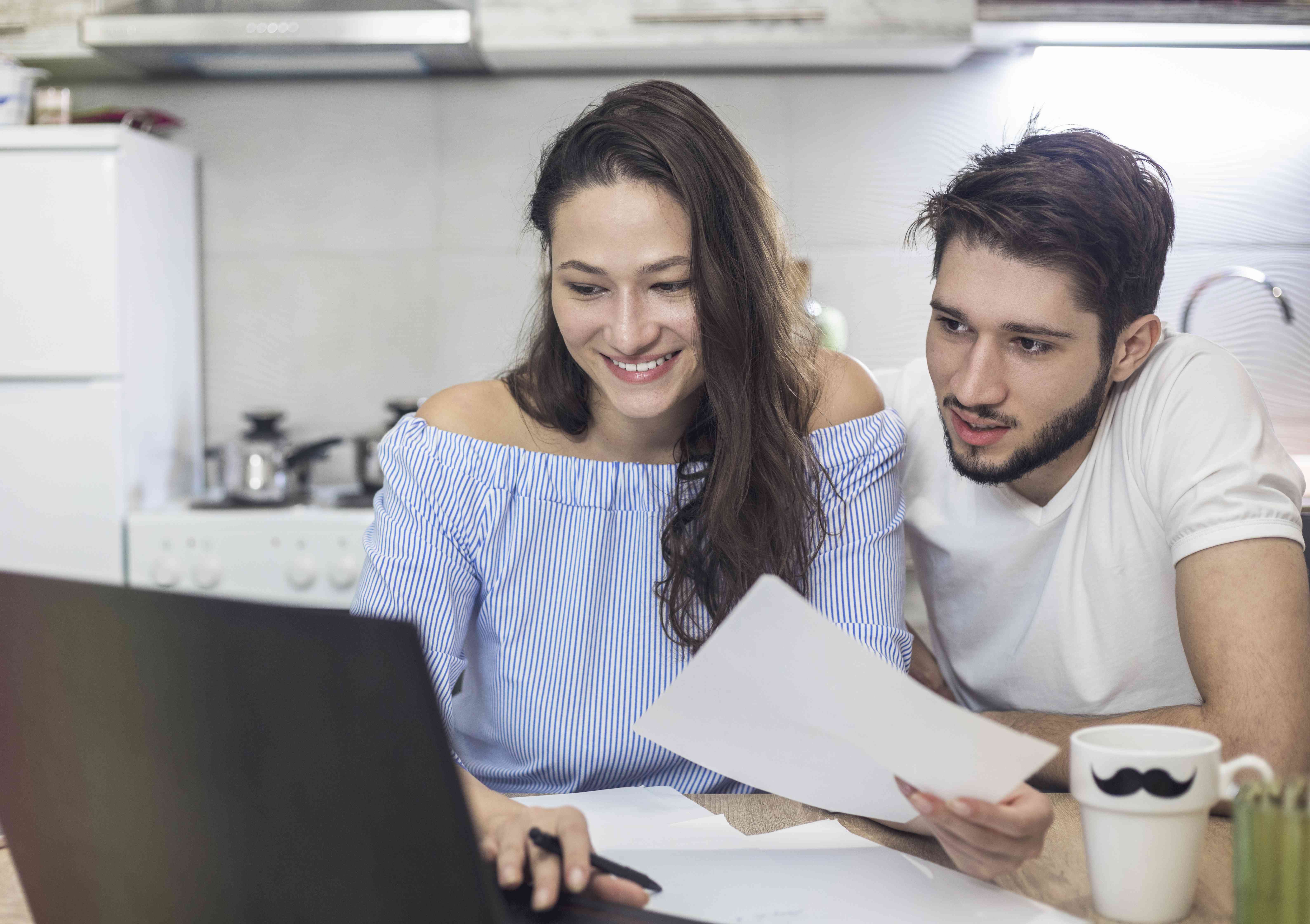 Young couple sitting at kitchen table and looking happily together at a laptop and financial documents