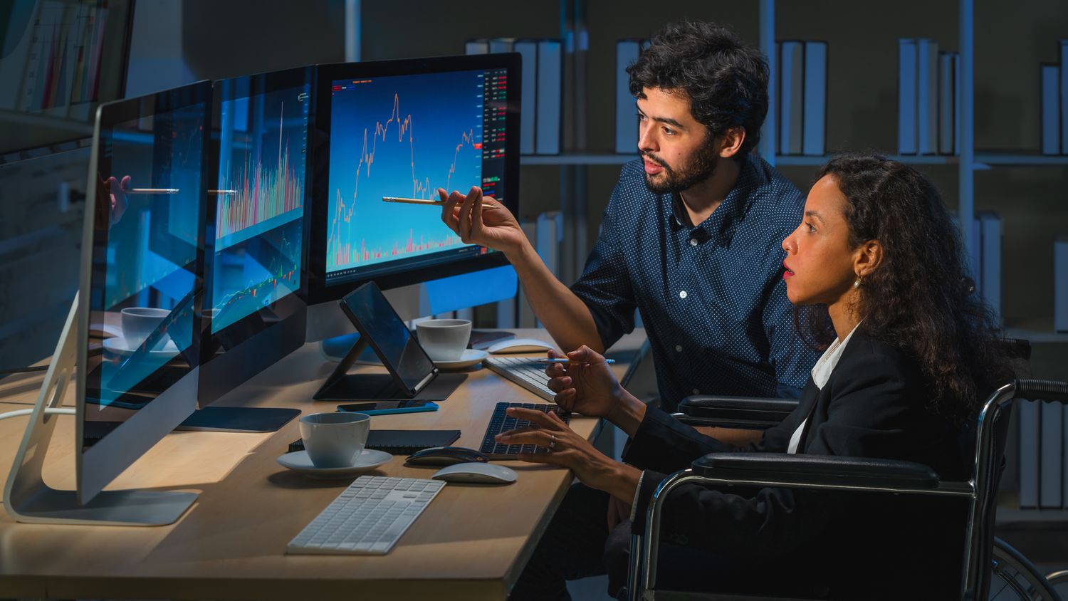 Two stock traders sit in a darken office analyzing price charts displayed on monitors to decide on pricing for future contracts.