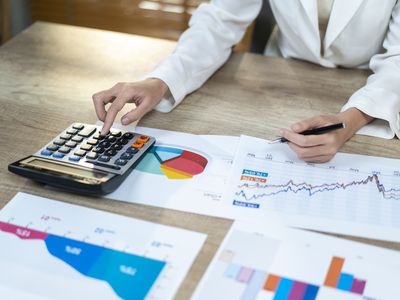 A close-up of a woman's hands using a calculator, while she sits at a desk with several documents displaying financial charts. 