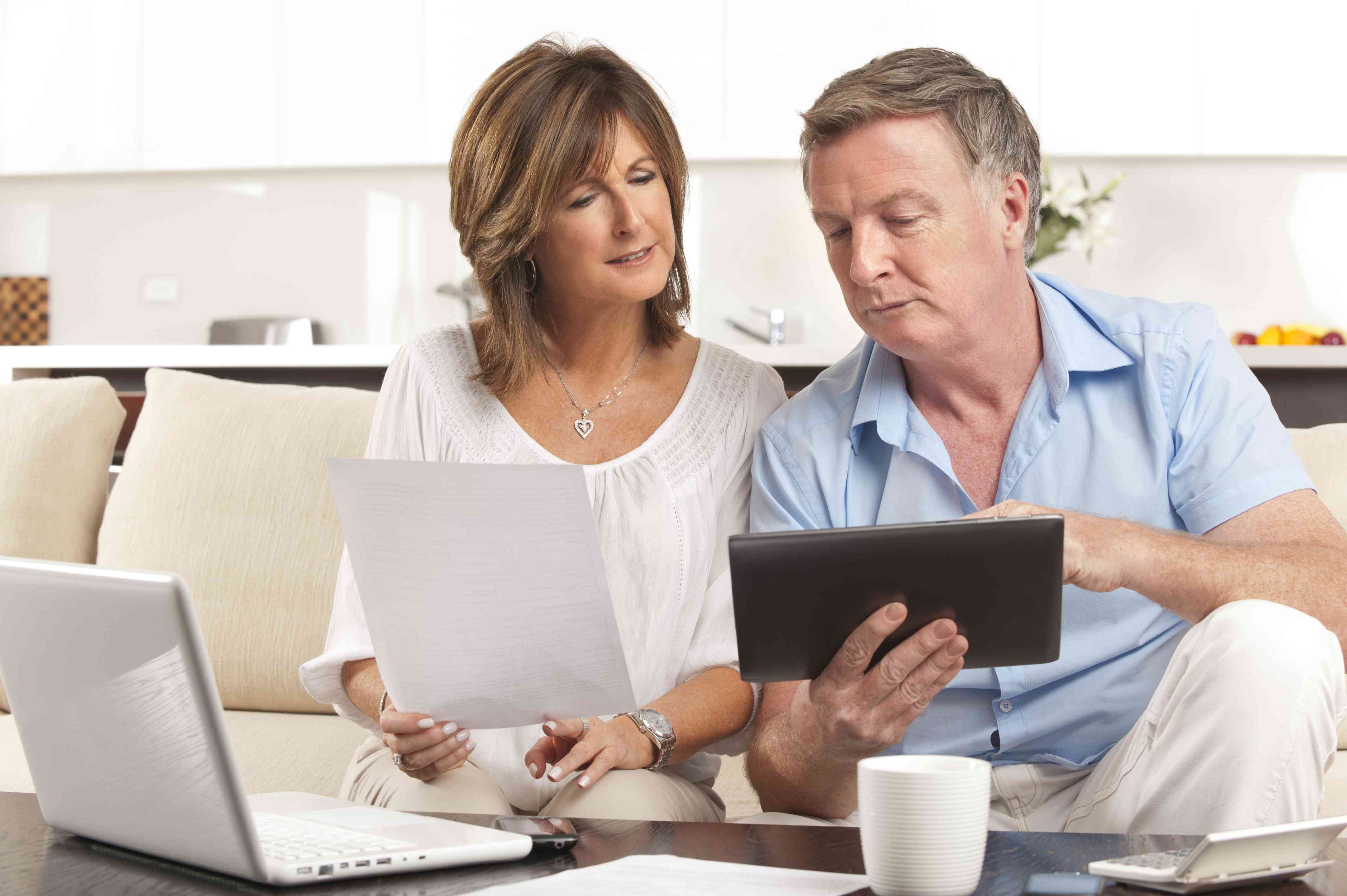 Older couple sitting on living room couch and looking together at a laptop, tablet, and documents