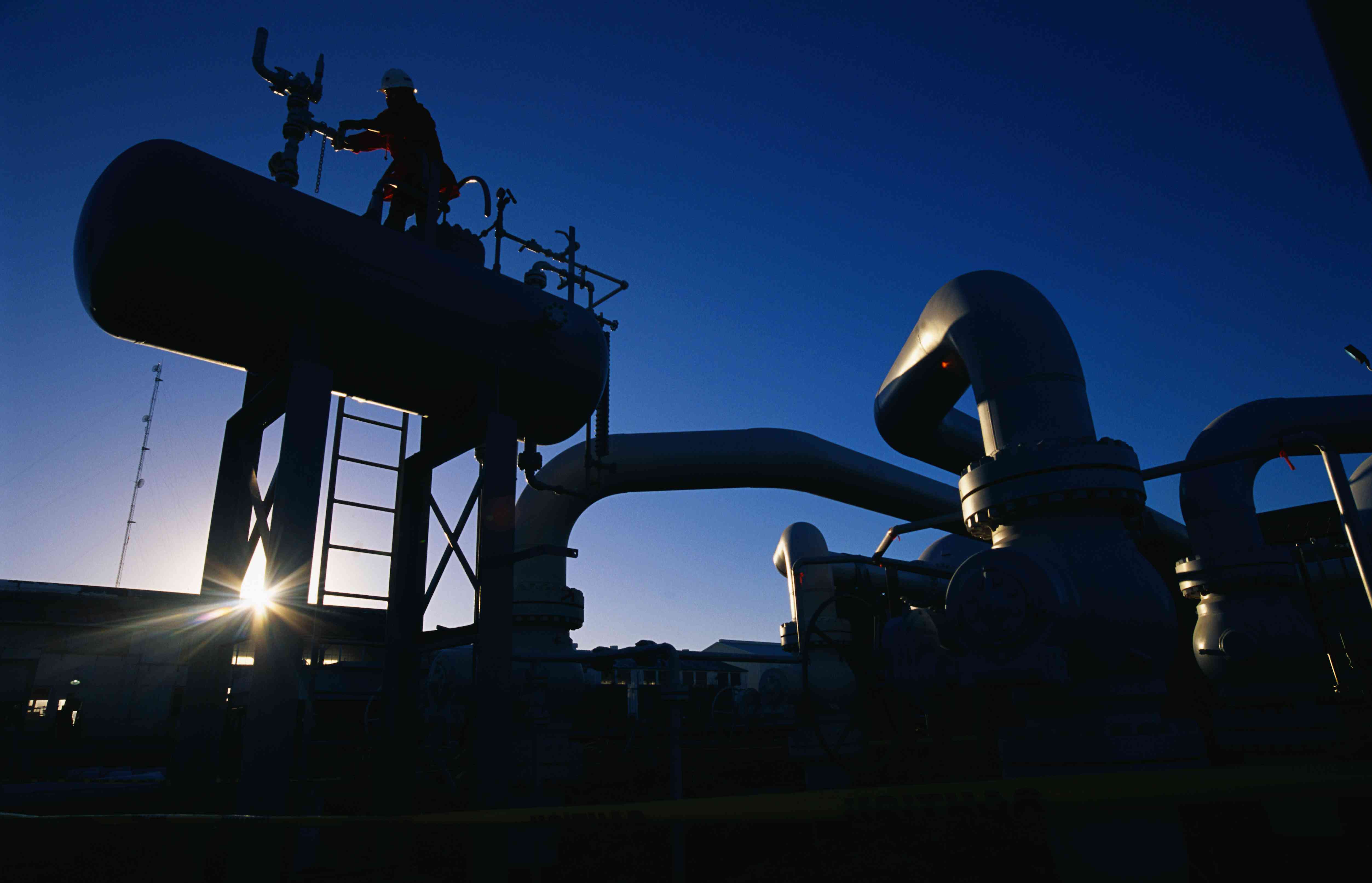 A natural gas production facility is silhouetted against the rising sun as a worker turns a valve on top of a large tank.