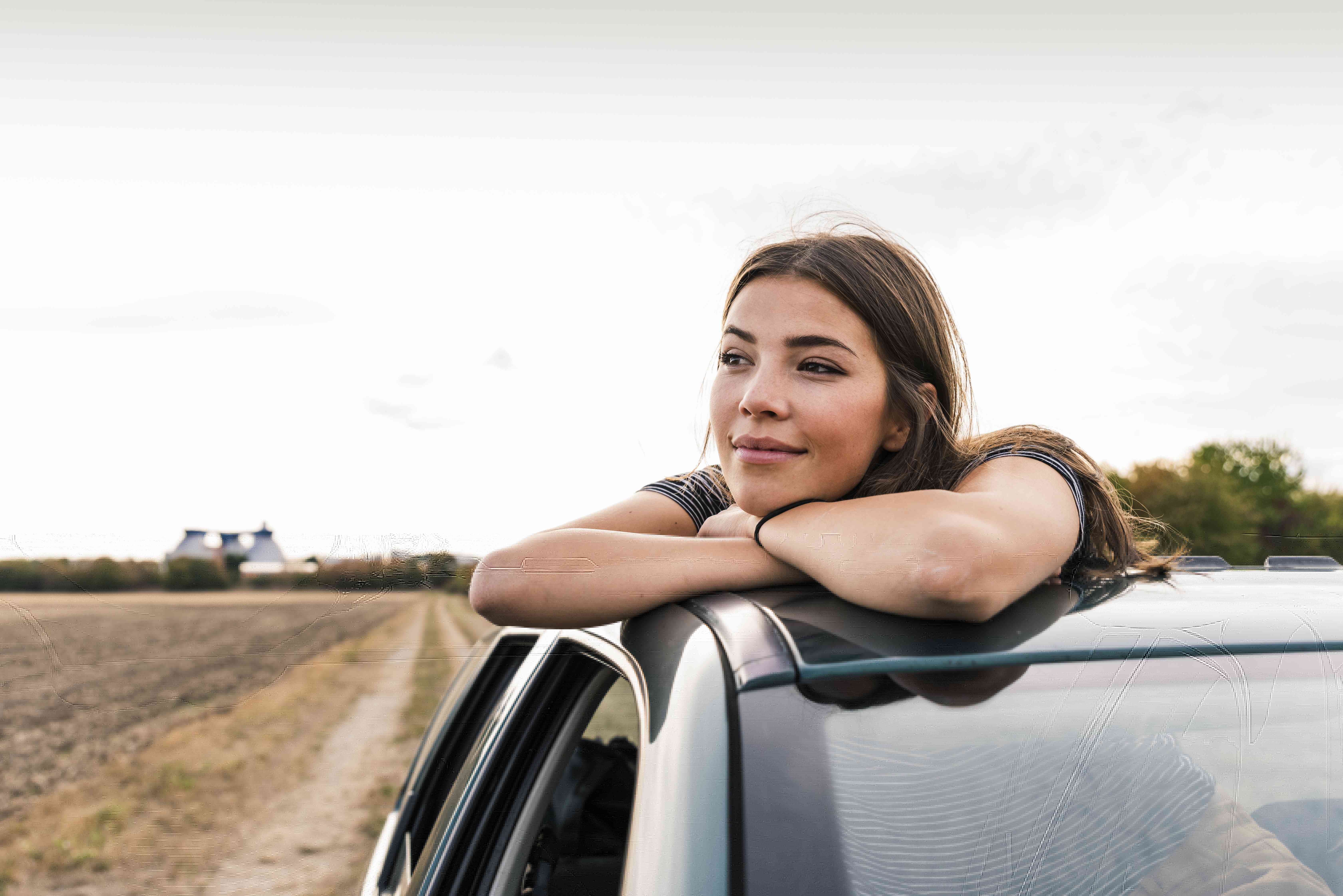 A young woman stands through a sunroof of a car.