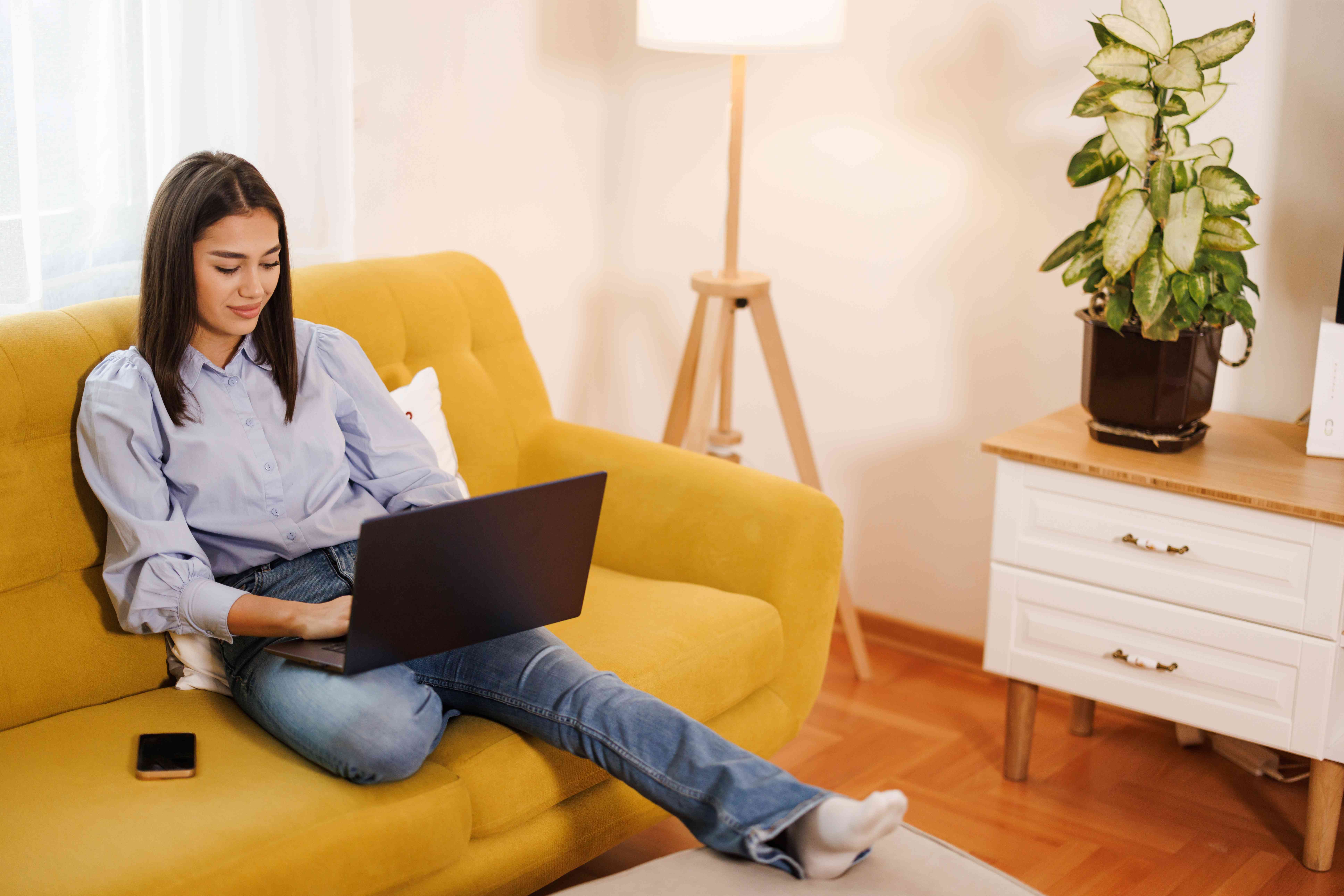 Young woman sitting on her living room couch and working on her laptop with a slight smile